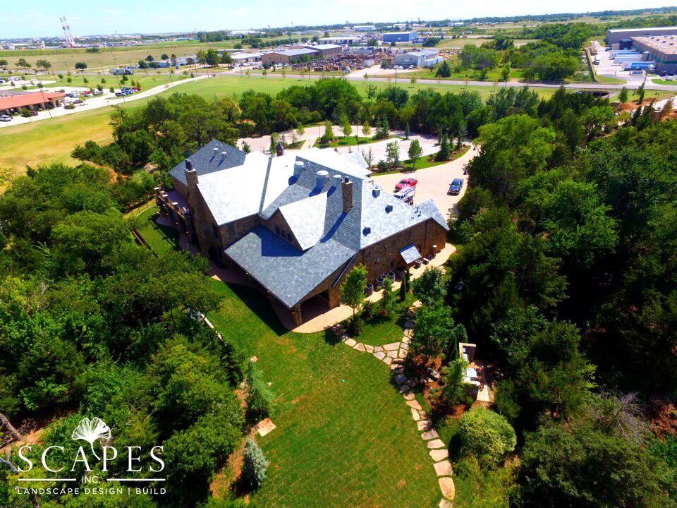 Aerial view of a stone building with a dark roof surrounded by green trees and landscaping; a grassy lawn leads to a stone pathway.
