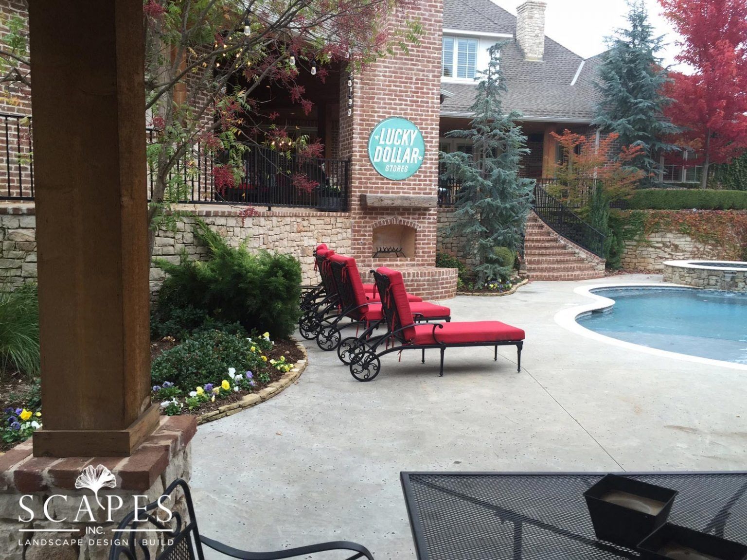 Patio with red lounge chairs near a pool, brick fireplace, and a large house in the background.