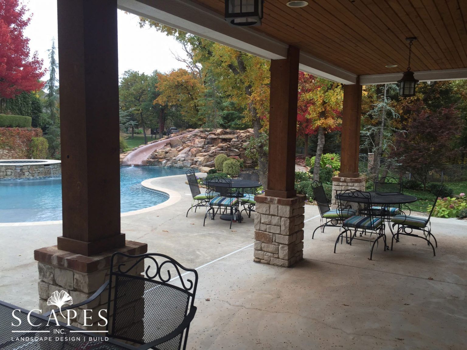 Patio with pool view. Brown columns with stone base, tables and chairs. Pool with waterfall and landscaping in background.