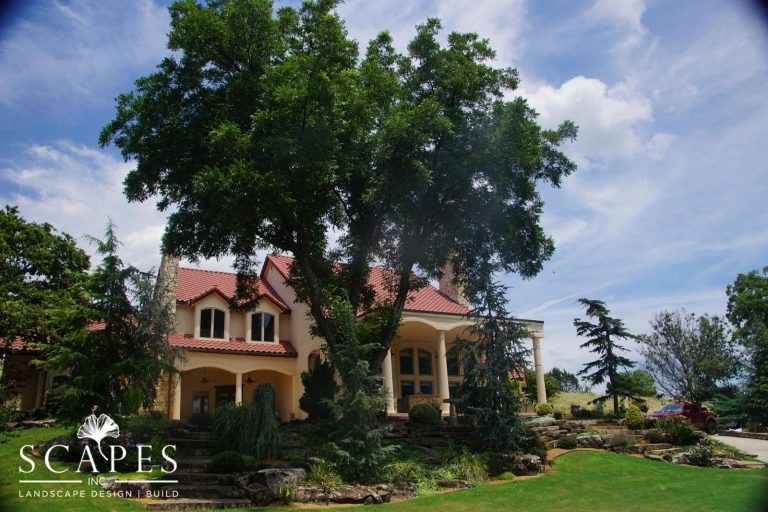 Beige house with red roof, large trees, and landscaped yard under a blue sky.