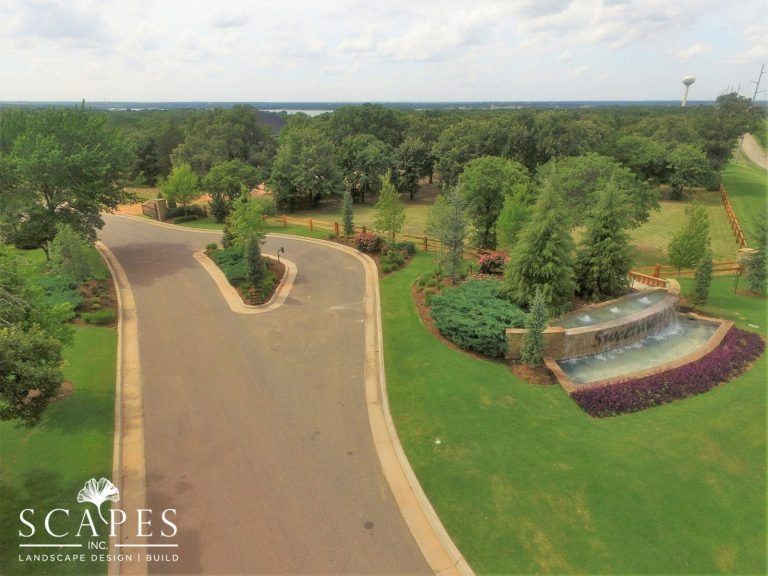 A residential street with a water fountain feature surrounded by green grass and trees under a cloudy sky.