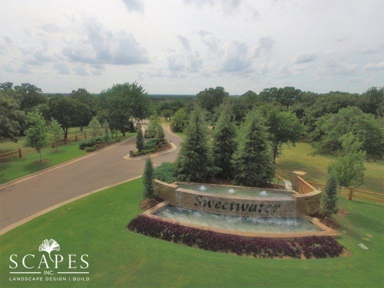A landscaped entrance to the Sweetwater neighborhood features a fountain, trees, and a brick sign under a cloudy sky.