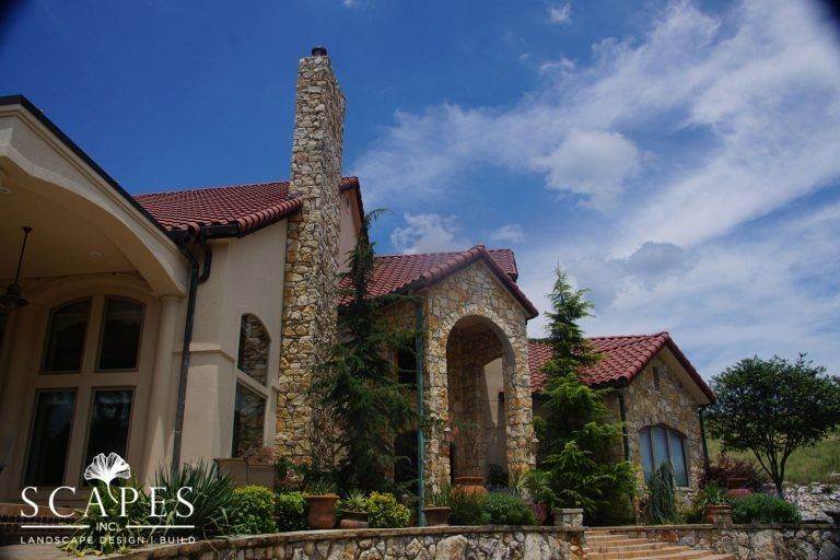 Stone house with red-tile roof, archway entrance, and tall chimney under a blue sky with clouds.