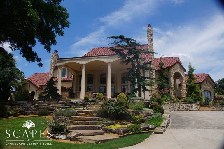 A large, luxurious home with a stone facade and a red tile roof. The home features a grand entrance with pillars, a landscaped front yard, and a winding driveway.