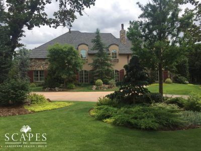 A large house with a brick and stucco facade, surrounded by green landscaping and trees. A circular driveway leads up to the house.