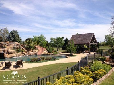 A luxury backyard with a pool, slide, and gazebo. The scene includes a grassy lawn, trees, and a black fence under a blue sky.