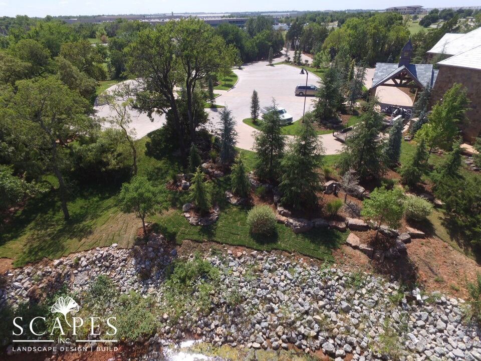 Aerial view of landscaped area with trees, rocks, and parking lot. Signage and a building with wooden accents are also visible.
