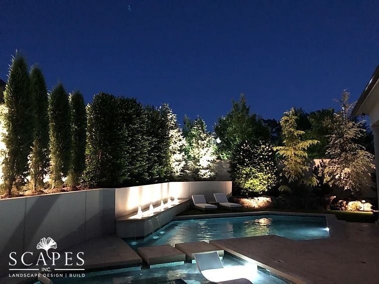 Nighttime outdoor scene: Illuminated pool with surrounding landscaping and trees. Low walls with water feature and lounge chairs under the dark sky.