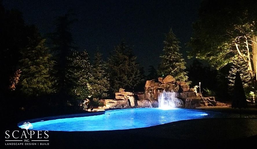 Night view of a backyard swimming pool with a waterfall feature illuminated with blue and white lights. Trees surround the pool.