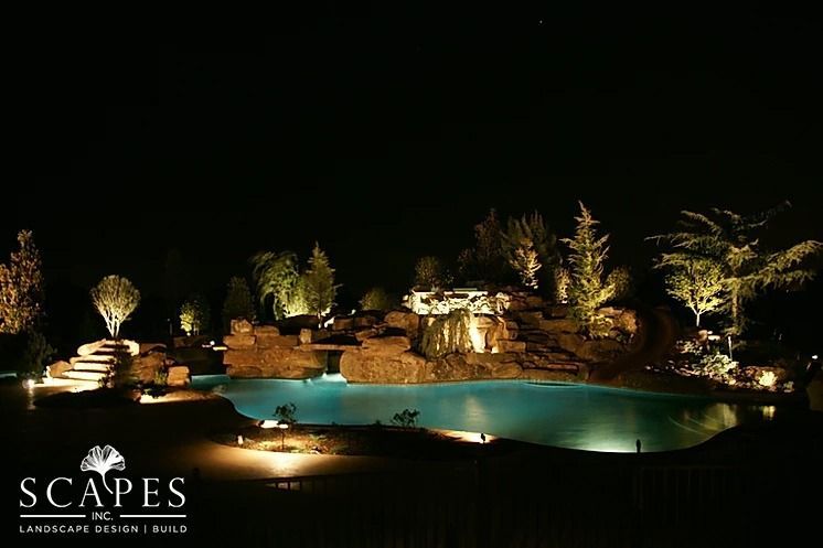 Nighttime view of a luxury backyard with pool, waterfall, and landscape lighting illuminating trees and rock formations.