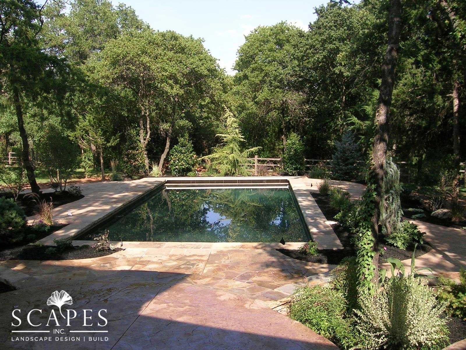 A rectangular swimming pool surrounded by stone patio in a wooded setting. Trees and foliage are abundant.