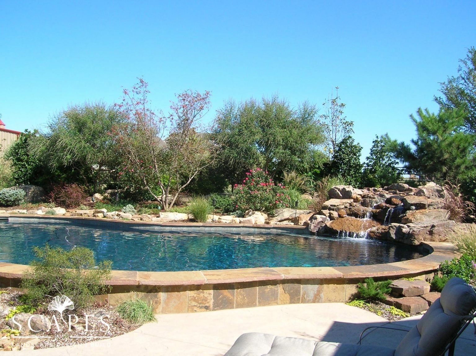 A backyard swimming pool with a waterfall feature surrounded by landscaping under a blue sky.