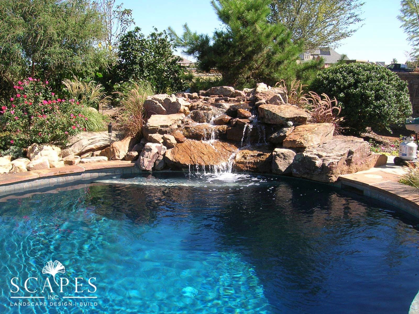 A waterfall cascades into a blue pool, surrounded by rocks and lush greenery on a sunny day.