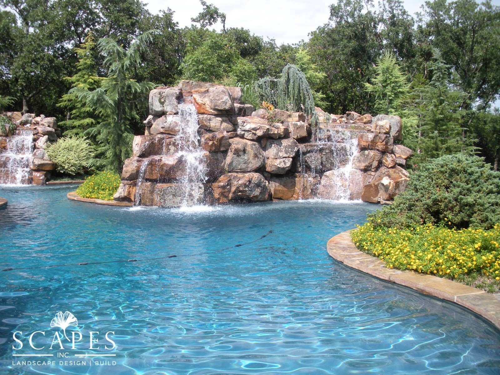 Swimming pool with waterfalls cascading over rocky features, surrounded by lush greenery and a stone border. The water is a vibrant turquoise.