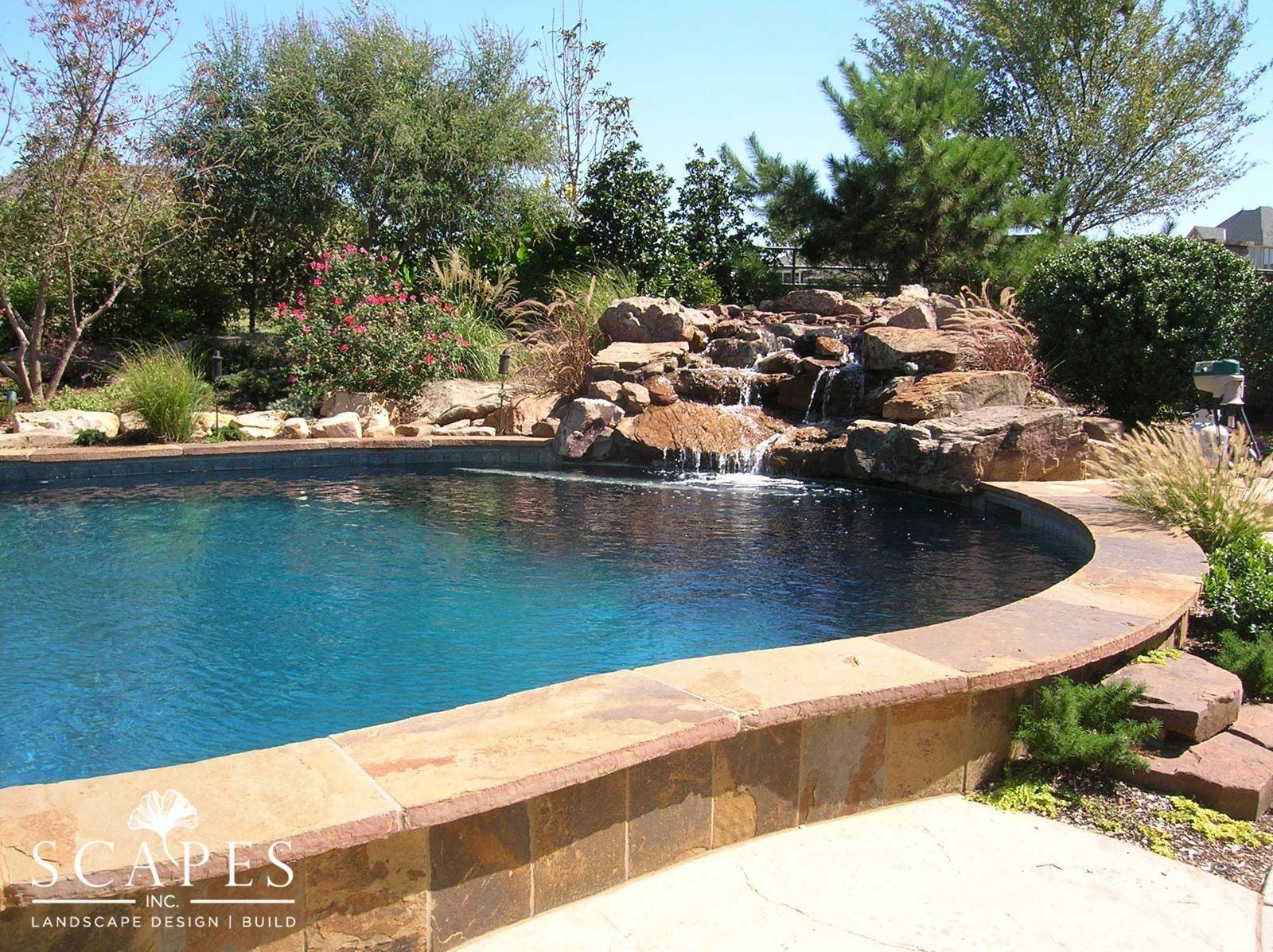 A swimming pool with a waterfall feature. The water is blue, surrounded by a stone border and lush greenery.
