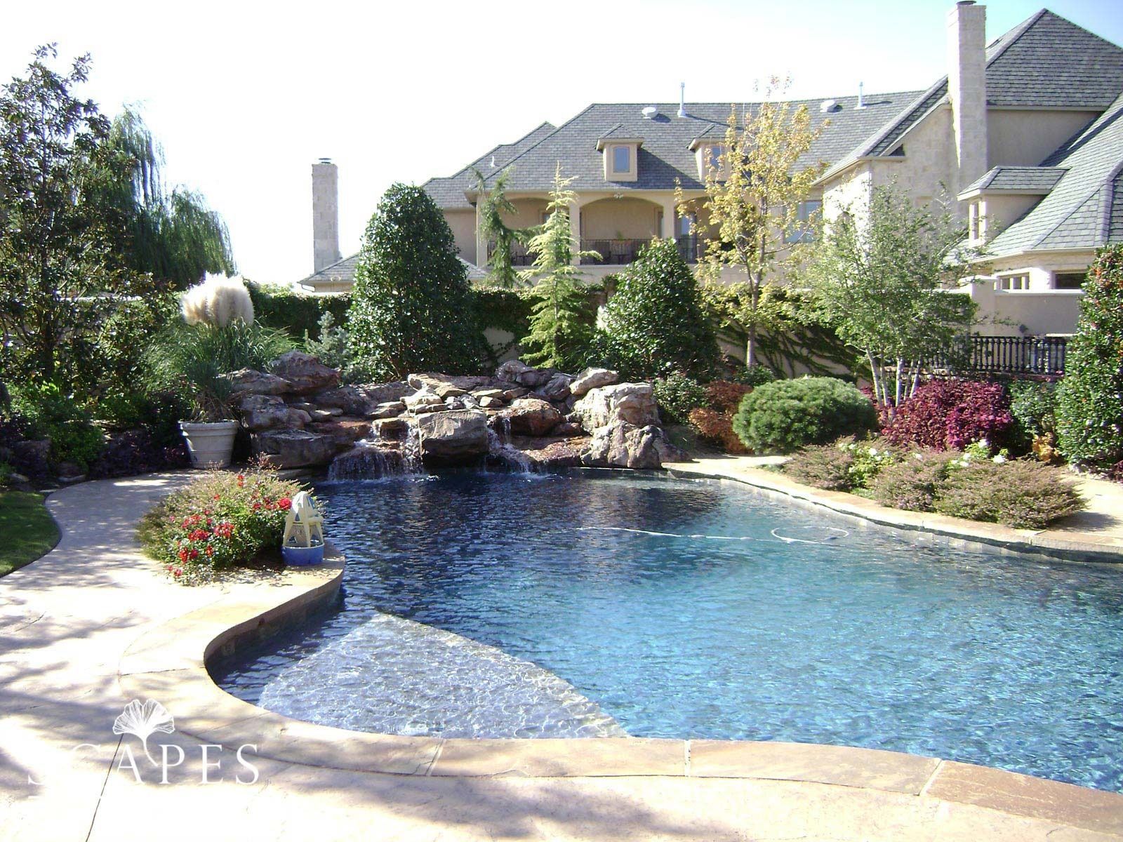Pool with waterfall feature in a landscaped backyard, stone patio and luxury home visible in the background.