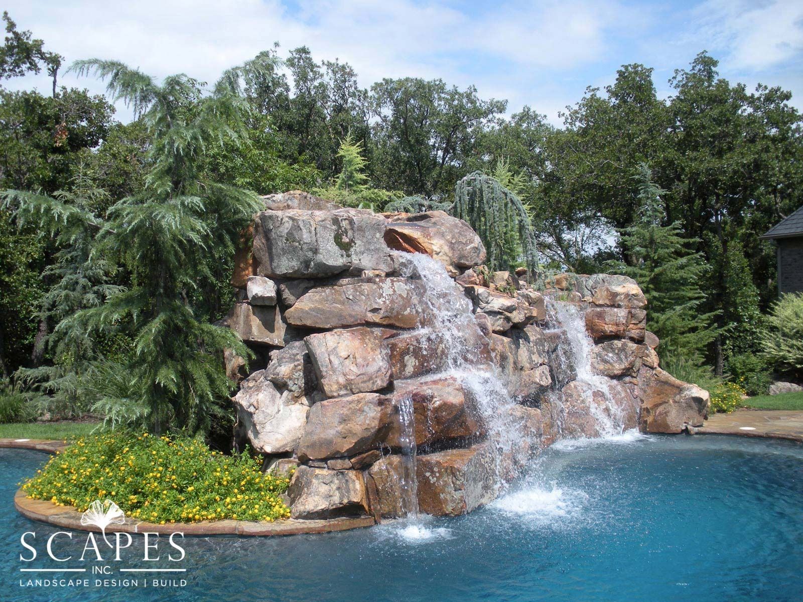 Waterfall feature cascading into a blue swimming pool, surrounded by greenery and rocks.