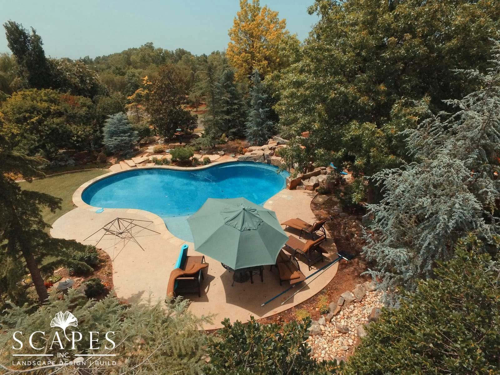 An aerial view of a backyard pool surrounded by lush trees and landscaping.  A patio features lounge chairs and a teal umbrella.