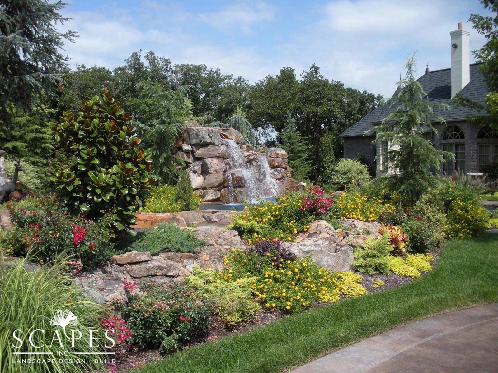 A landscaped backyard features a waterfall, lush greenery, and colorful flowers. A house with a stone facade is in the background.