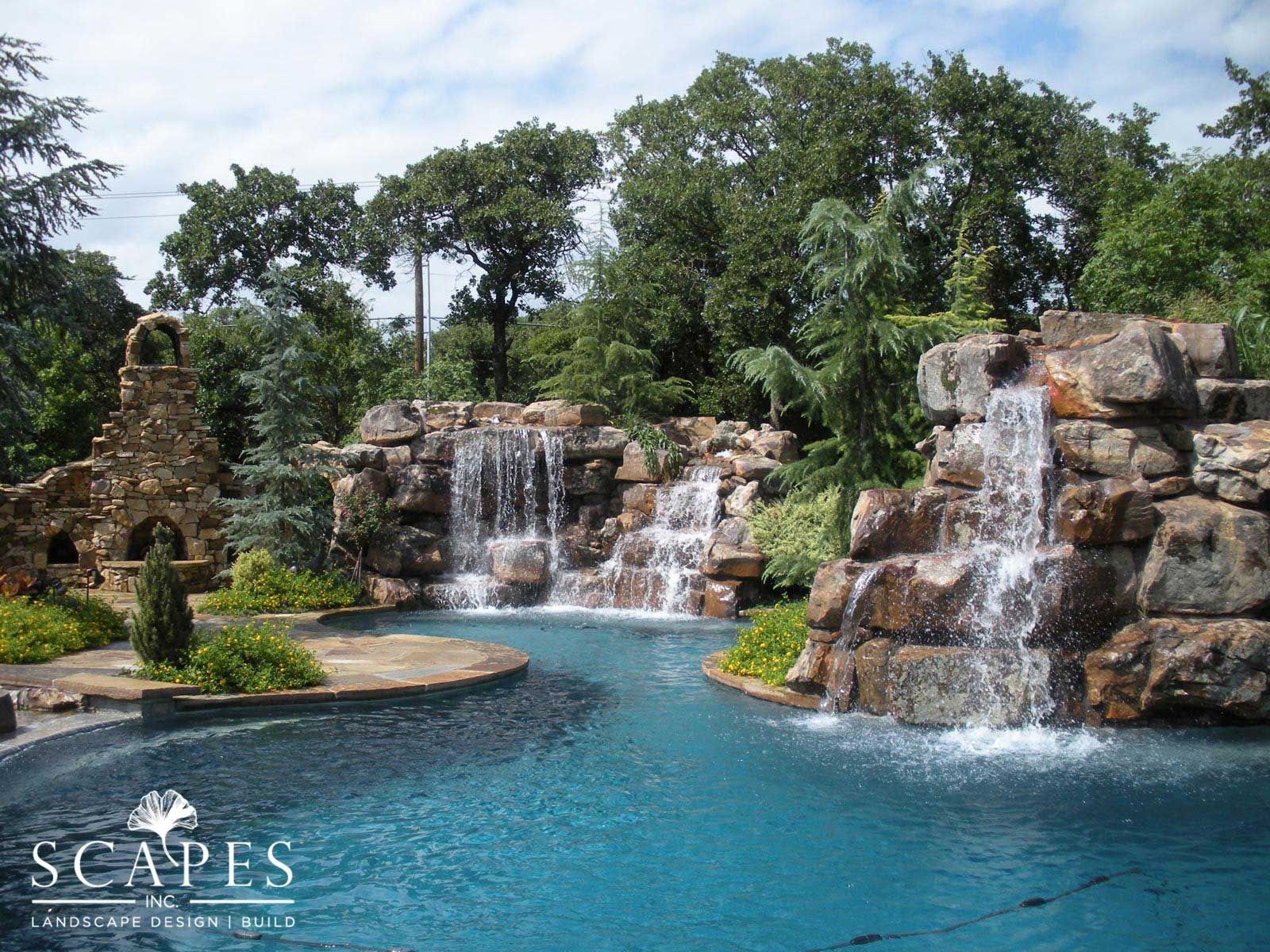 A swimming pool with two waterfalls cascading over rock formations, surrounded by lush greenery.