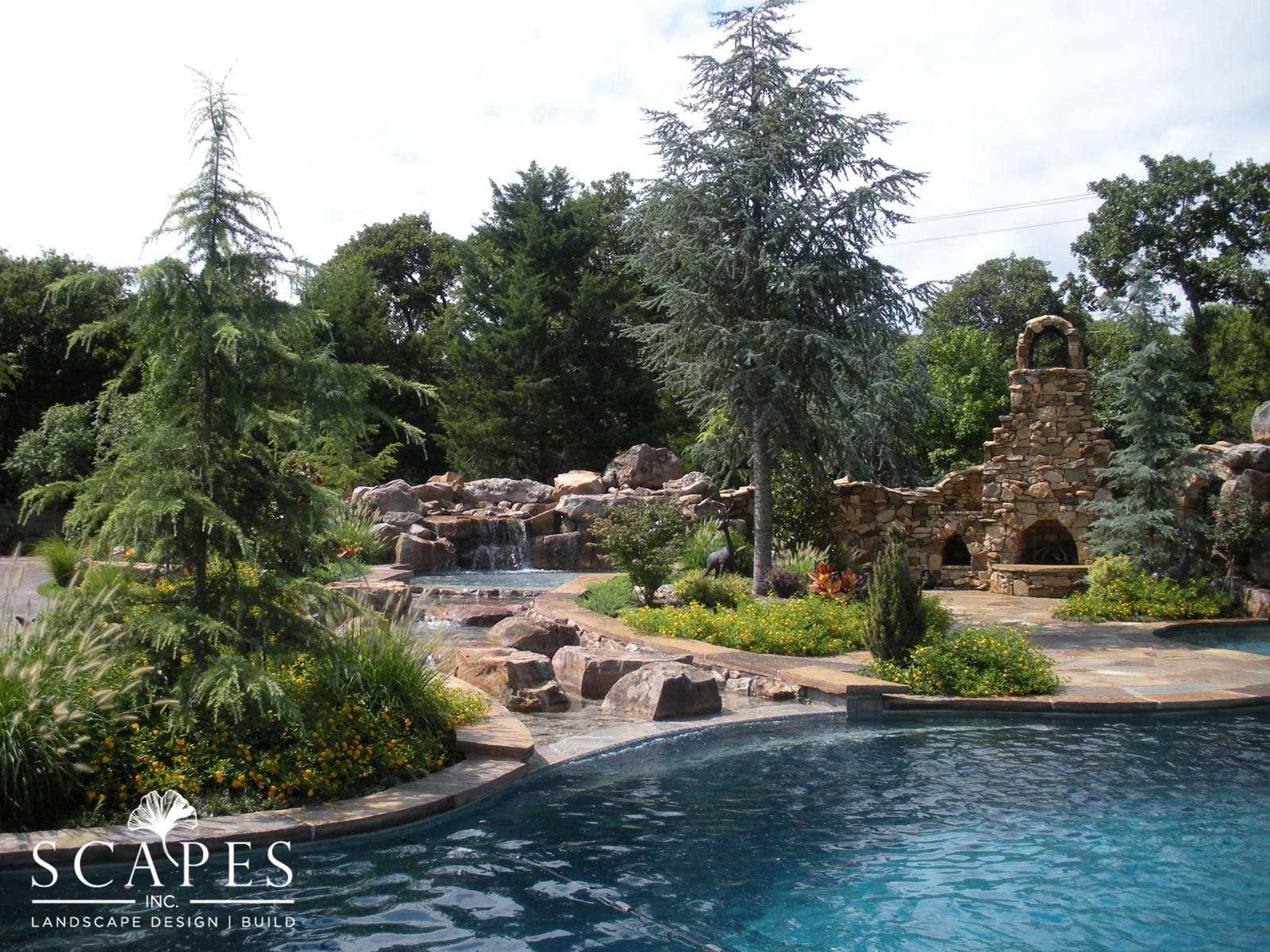 A backyard pool with a rocky waterfall feature, fireplace, and lush landscaping. Blue water and green foliage dominate the scene.