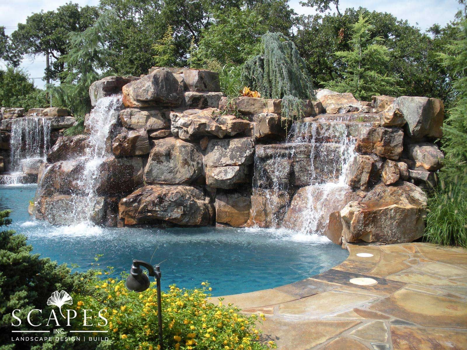 Waterfalls cascade into a blue pool, surrounded by brown rock and lush greenery.