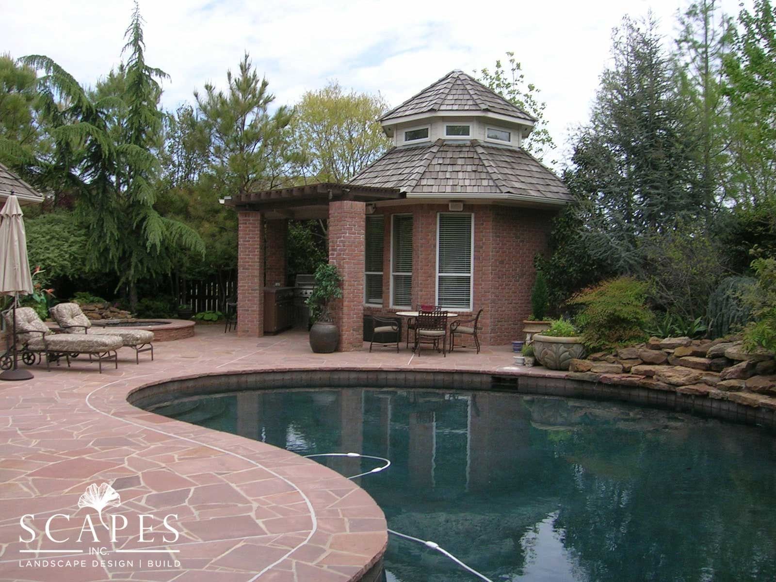 A pool area with a brick cabana, surrounded by landscaping. The cabana has a round roof and windows, and there is a seating area by the pool.