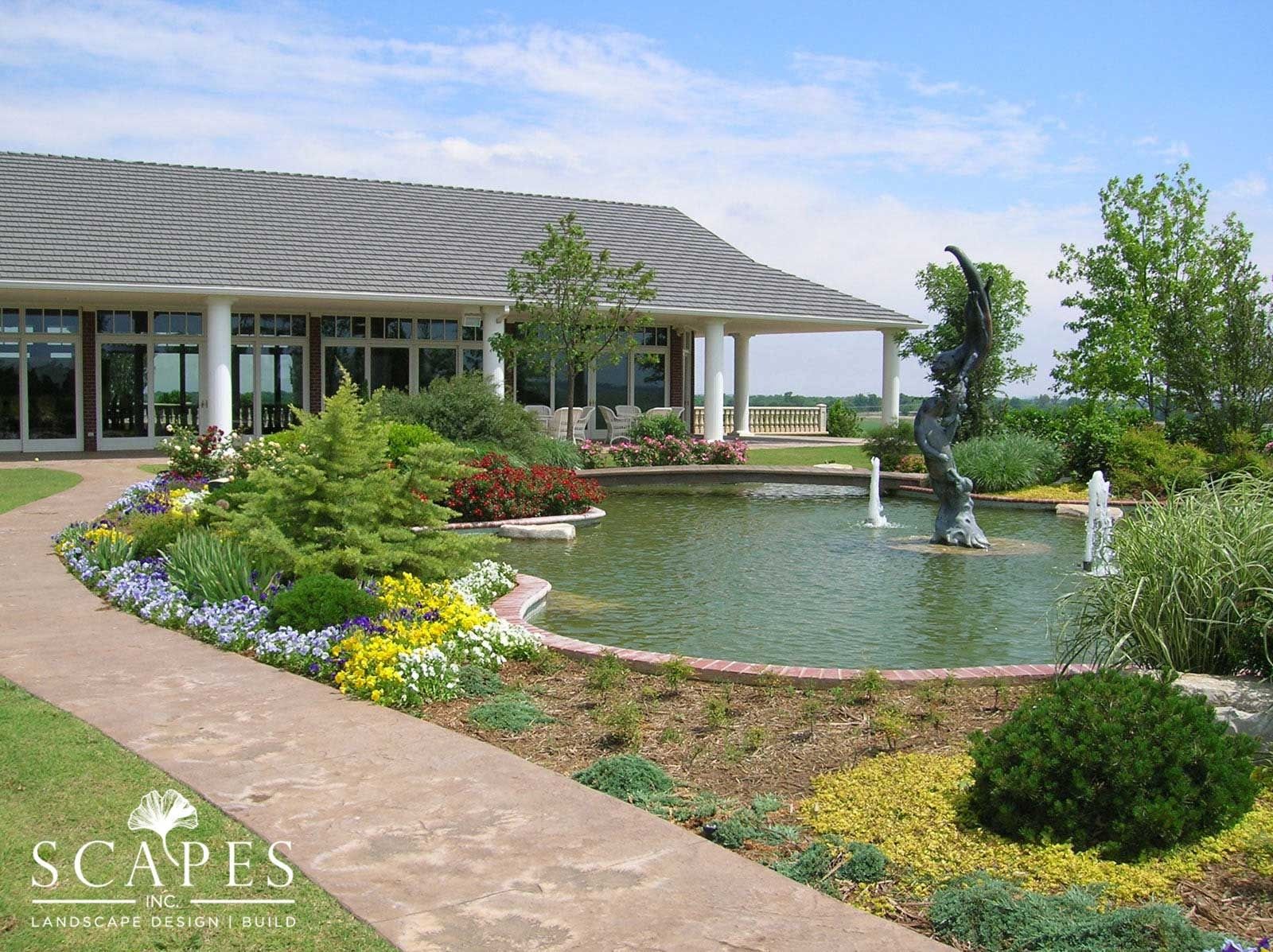 A paved path leads past a pond with a fountain and sculpture, towards a building with a covered porch.