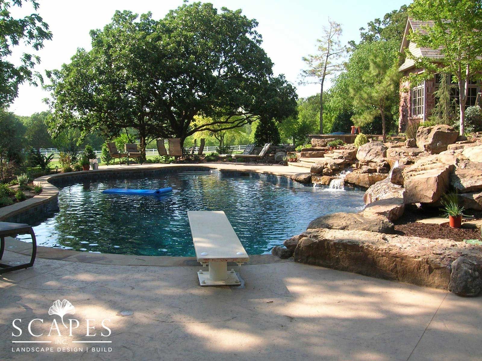 A backyard pool with a diving board, surrounded by stone features, greenery, and a house.