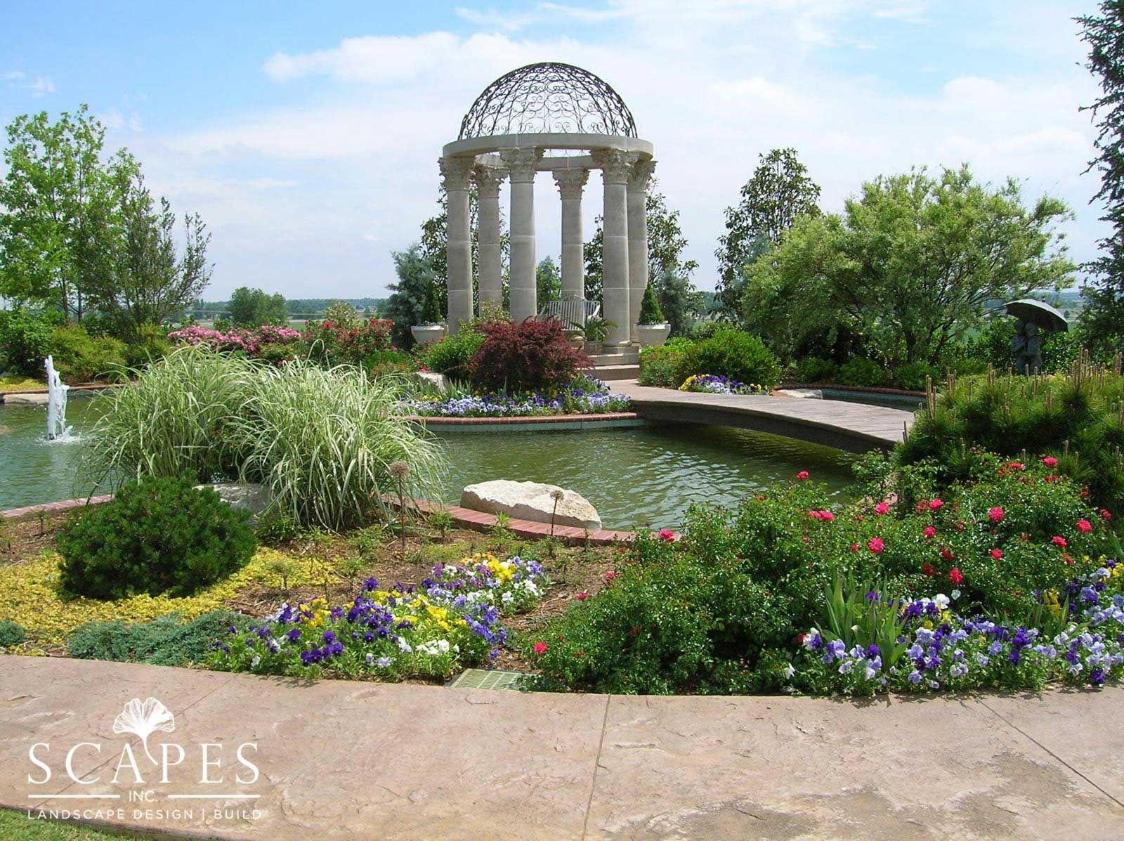 Stone gazebo with a domed top overlooks a pond and flowerbeds, with trees in the background.