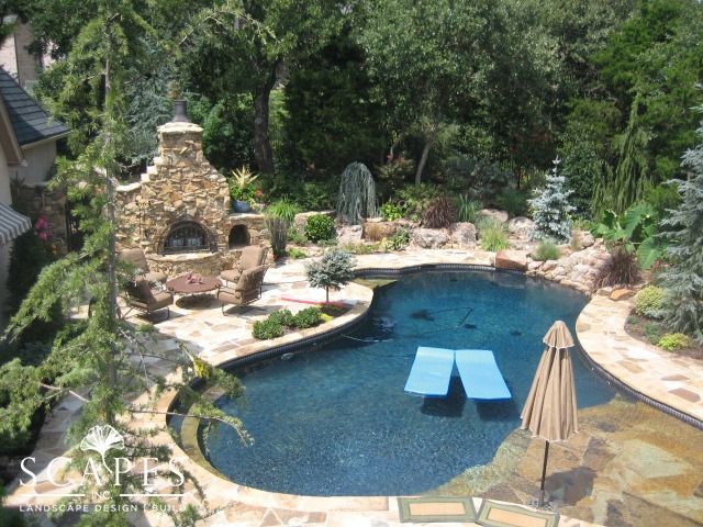 Outdoor pool with stone fireplace and seating area surrounded by lush landscaping. Two blue pool floats are in the water.