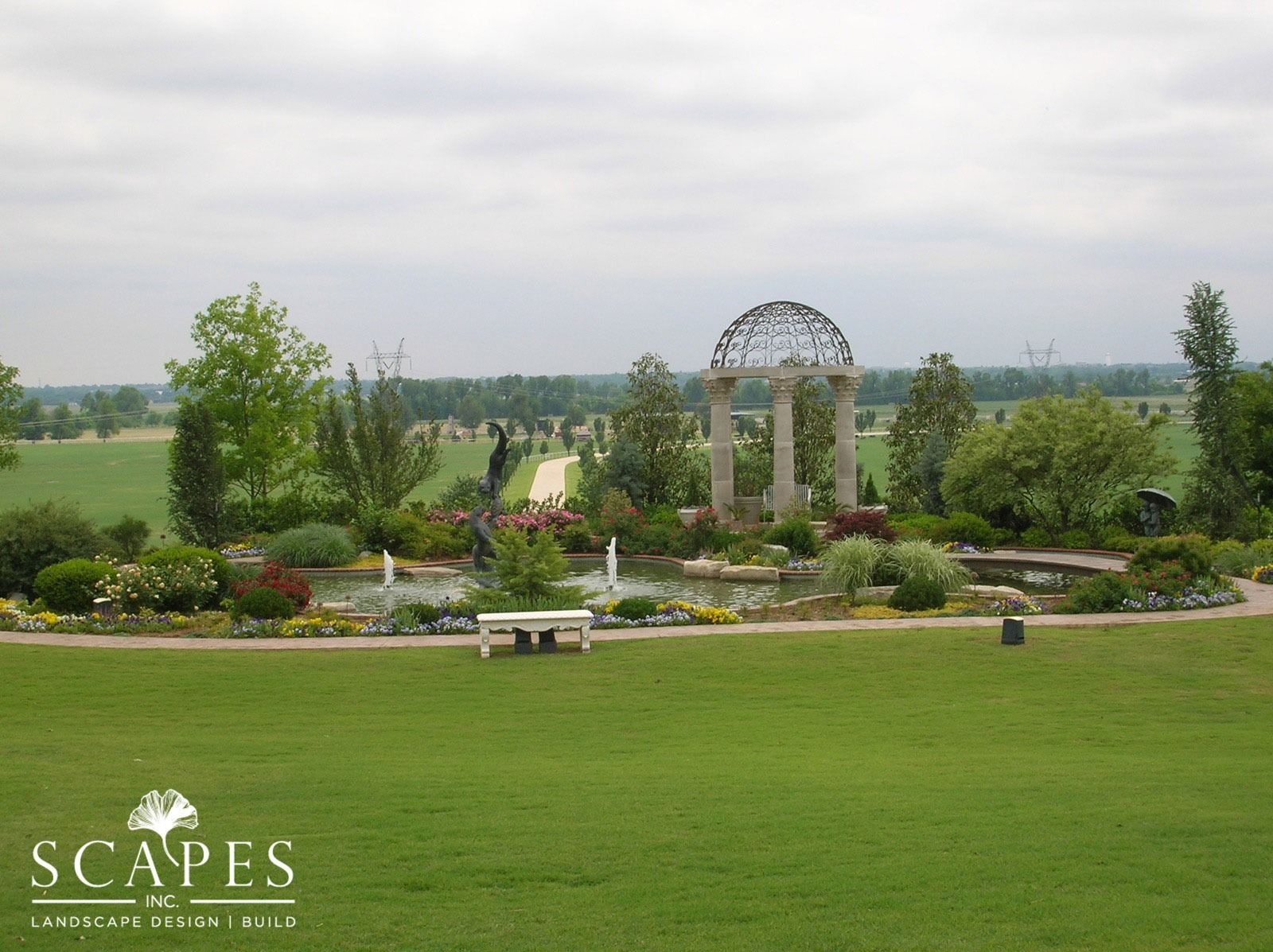 A grassy park with a gazebo, a pond, and a bench. Trees and fields are in the background under a cloudy sky.