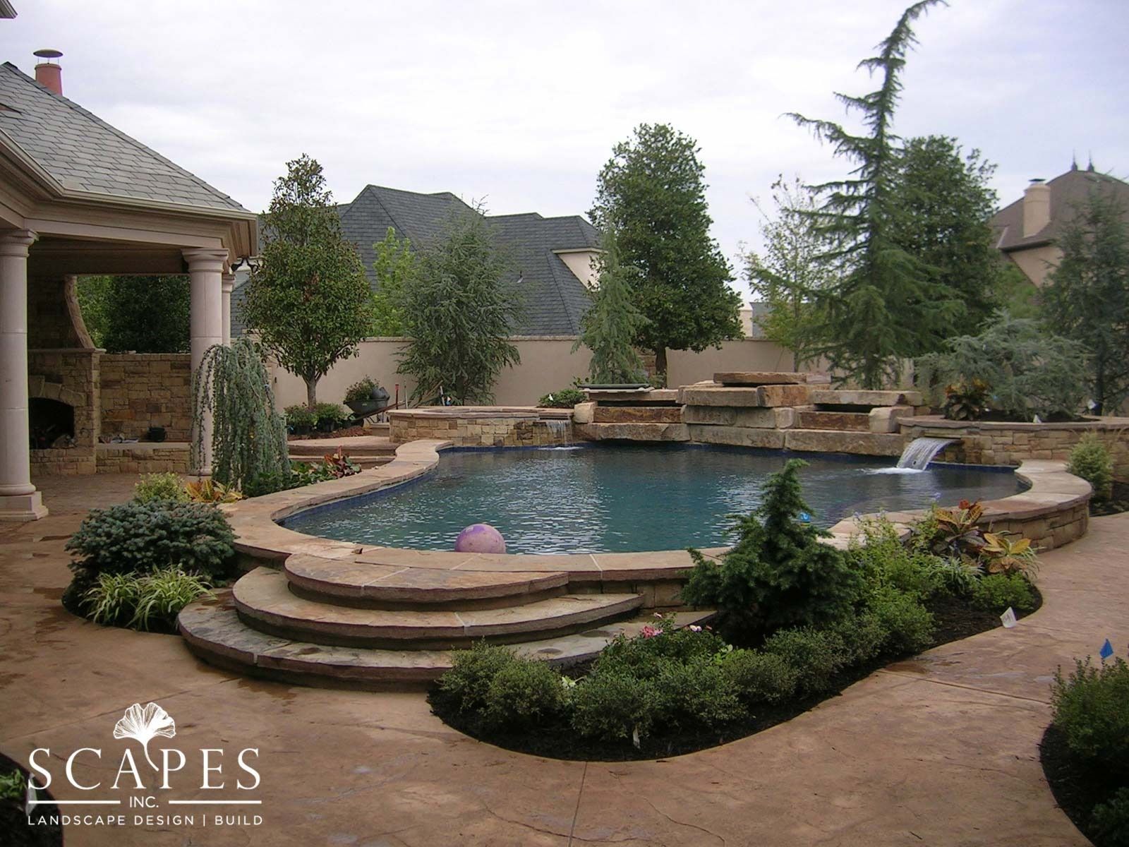 A backyard pool with stone coping, waterfalls, and landscaping. Includes a stone patio and a covered patio area.