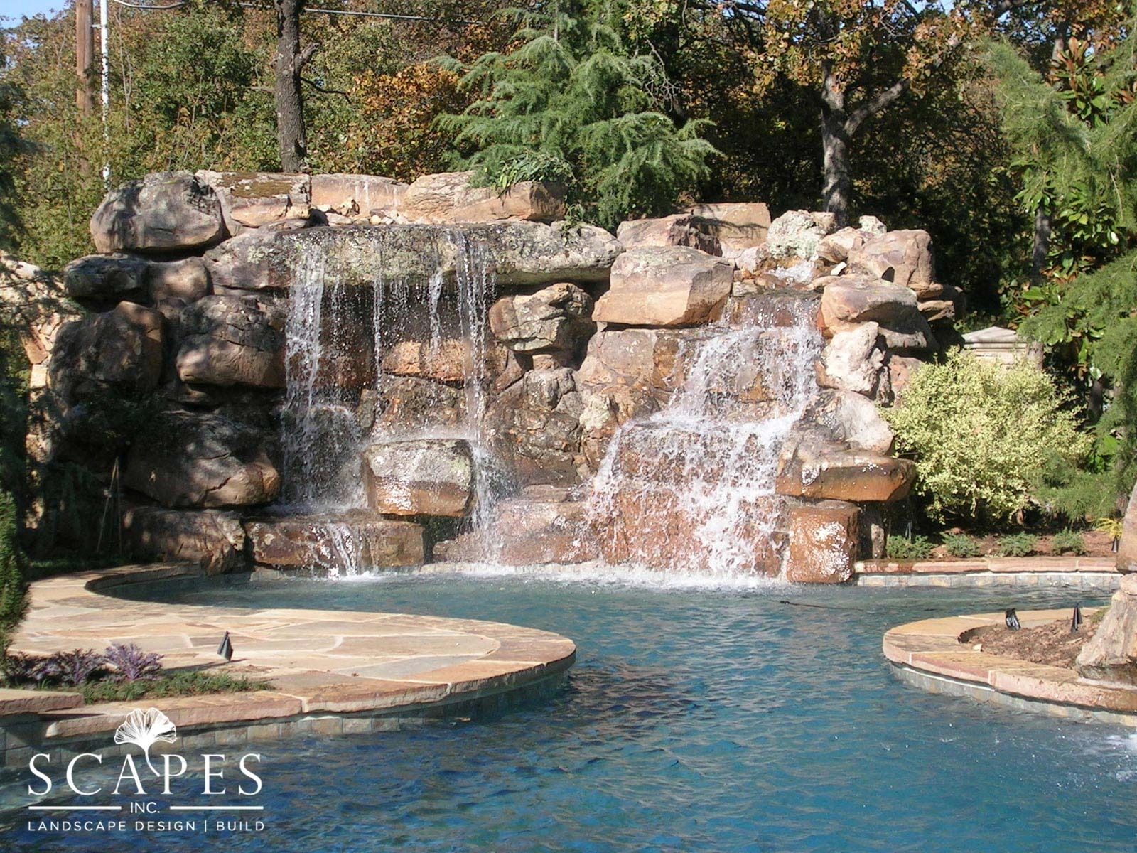 Waterfall cascading into a blue pool, surrounded by stone and greenery; natural outdoor setting.