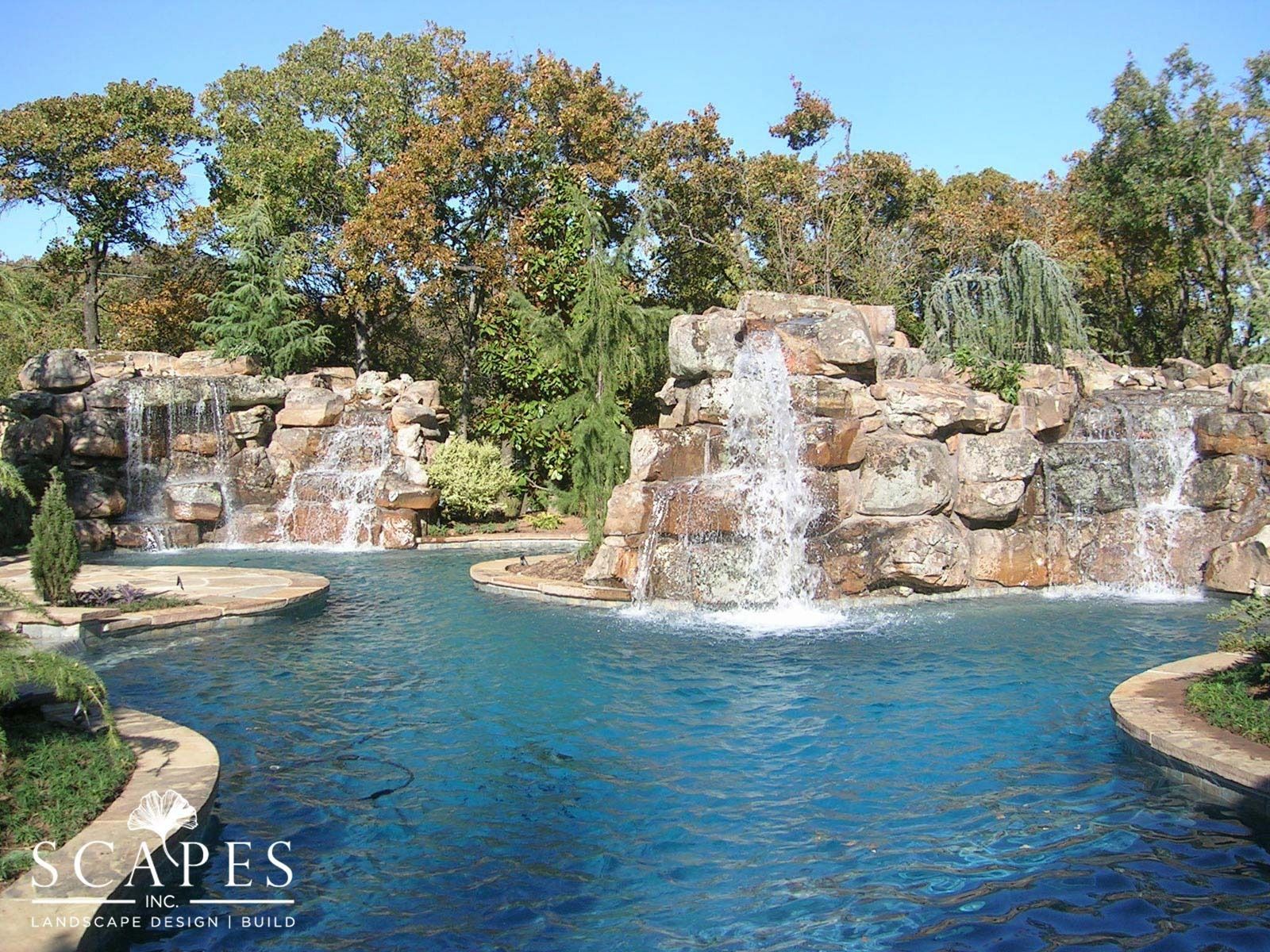 Two waterfalls cascade into a bright blue pool surrounded by rocky formations and trees under a clear sky.