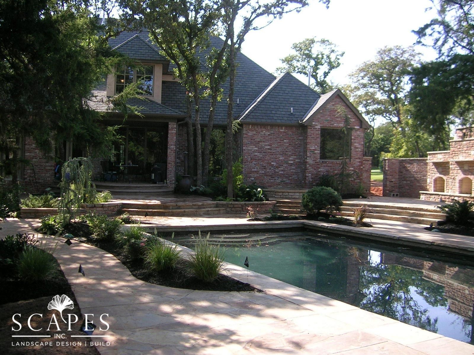 Backyard with a pool, brick house, and landscaping. The pool is dark blue, and the house is brown brick with a dark roof.
