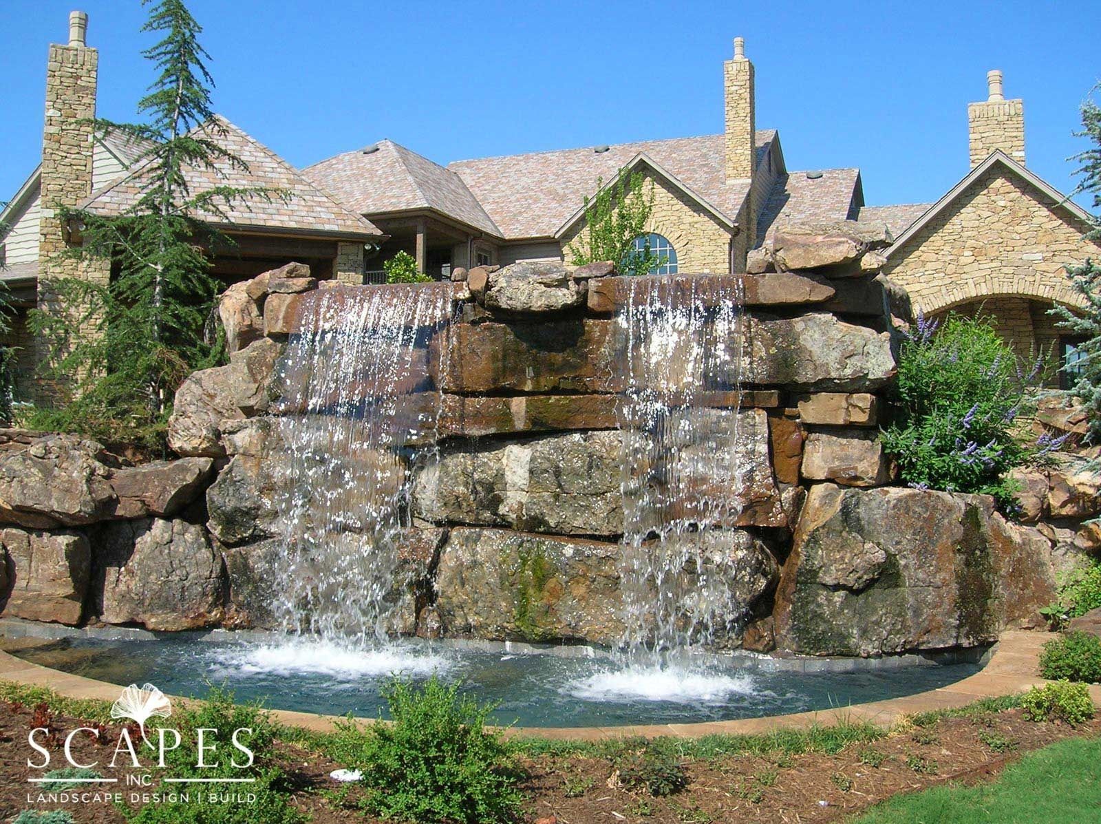 A large stone waterfall cascades into a pool, set in front of a multi-gabled stone house on a sunny day.