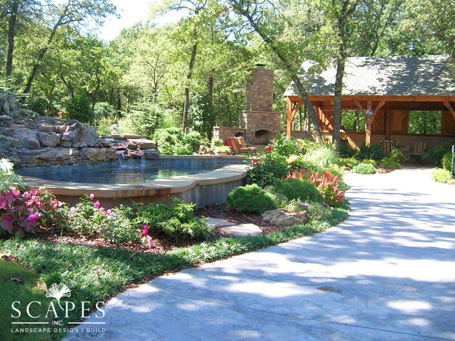 A stone patio and pool area with a waterfall, surrounded by greenery, a wooden shelter, and a stone fireplace under a sunny sky.