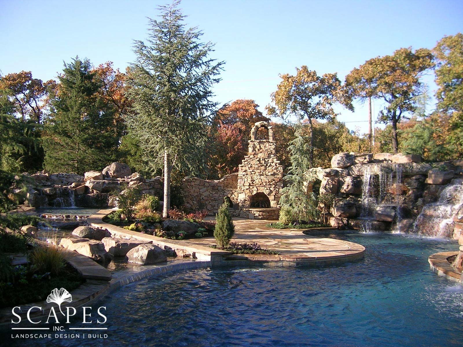 A large, elaborate outdoor pool area with waterfalls, rock formations, and a stone structure under a clear sky.