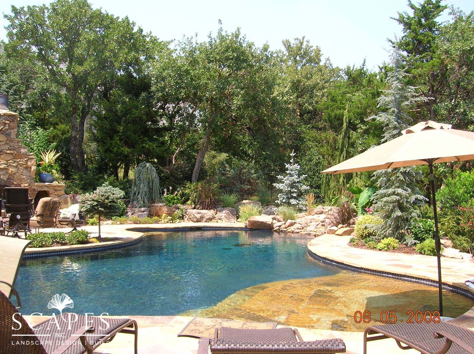 A backyard with a dark blue pool, rocky edge, trees, and an umbrella. Two lounge chairs are in the foreground.