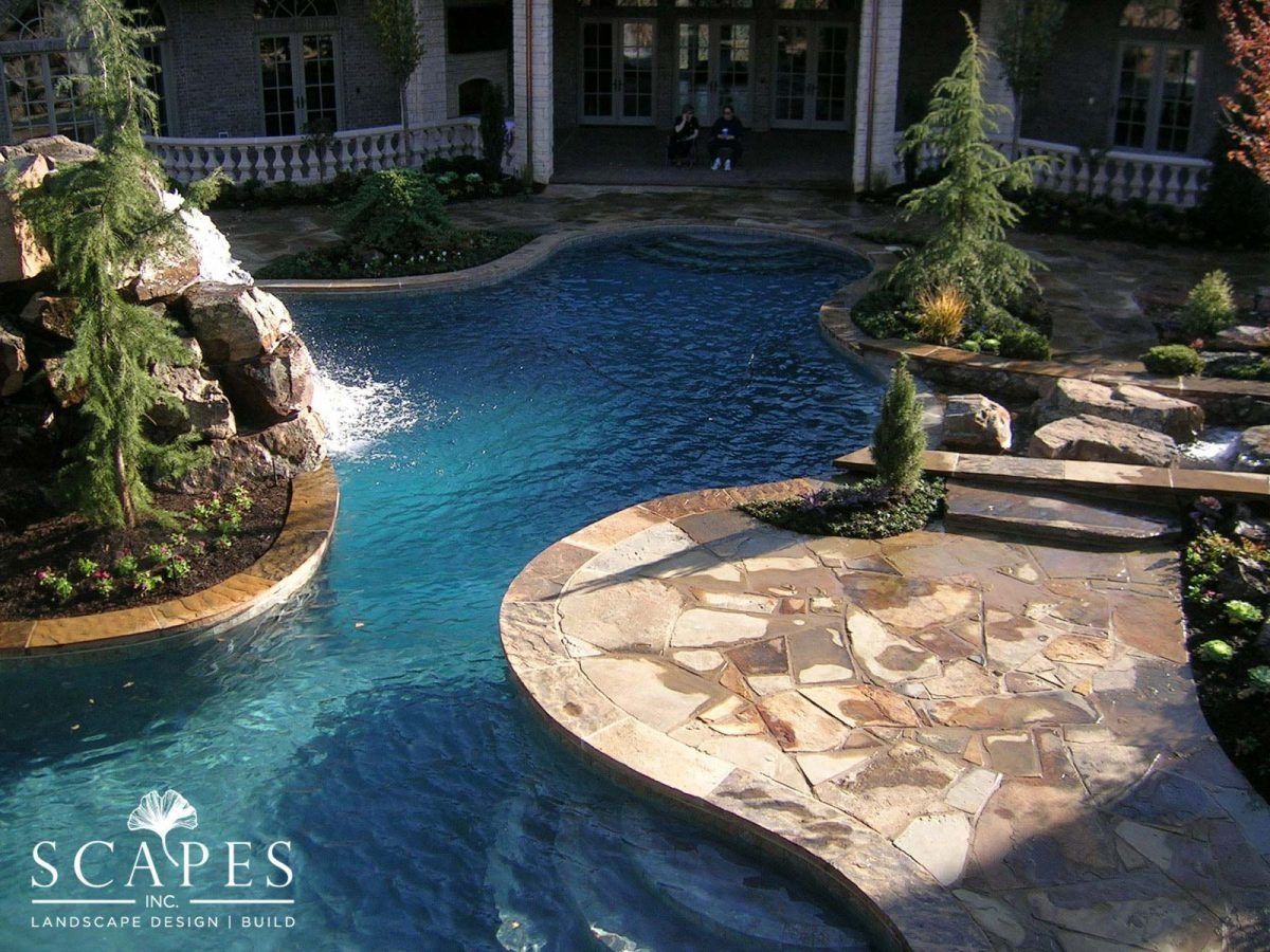 A luxurious pool with a waterfall, natural stone walkways, and lush landscaping in front of a large home.