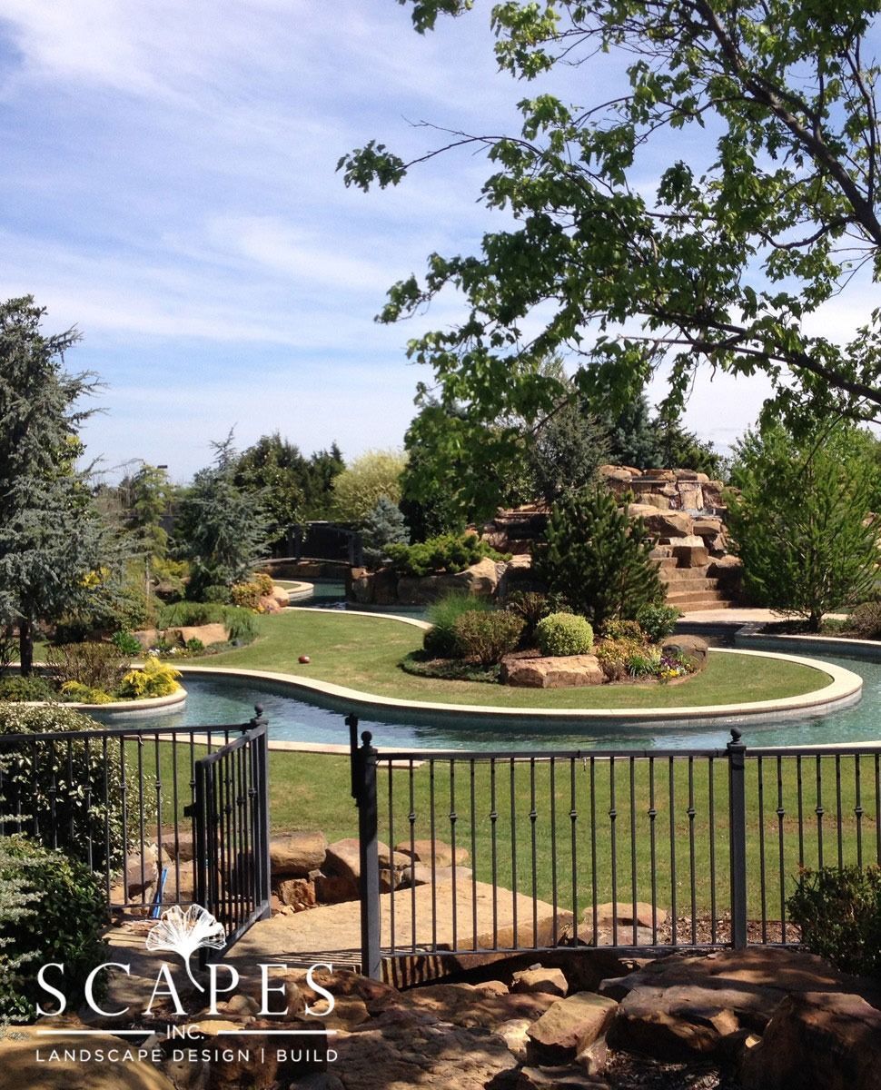 A landscaped garden with a water feature, lush greenery, and a rock waterfall, viewed through a black fence.