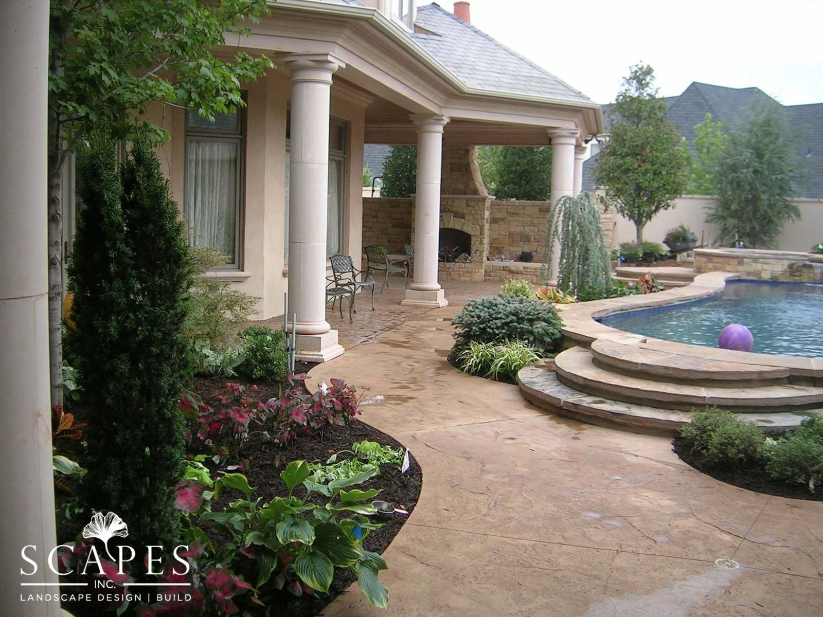 A luxury backyard with a pool, patio, and covered seating area featuring columns, plants, and stone work.