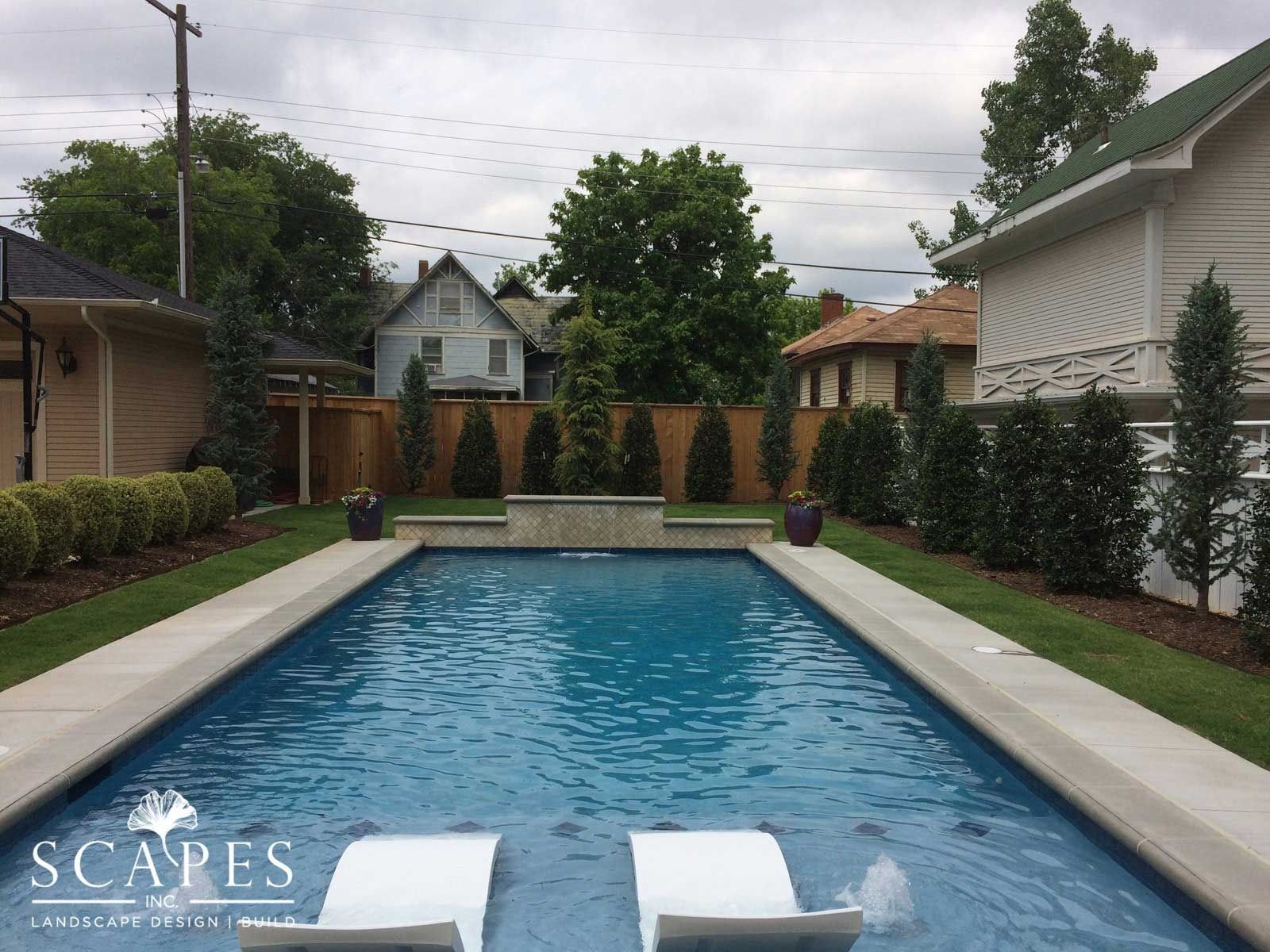 A rectangular pool with lounge chairs faces a privacy fence and homes in the background. Green plants border the pool and lawn.