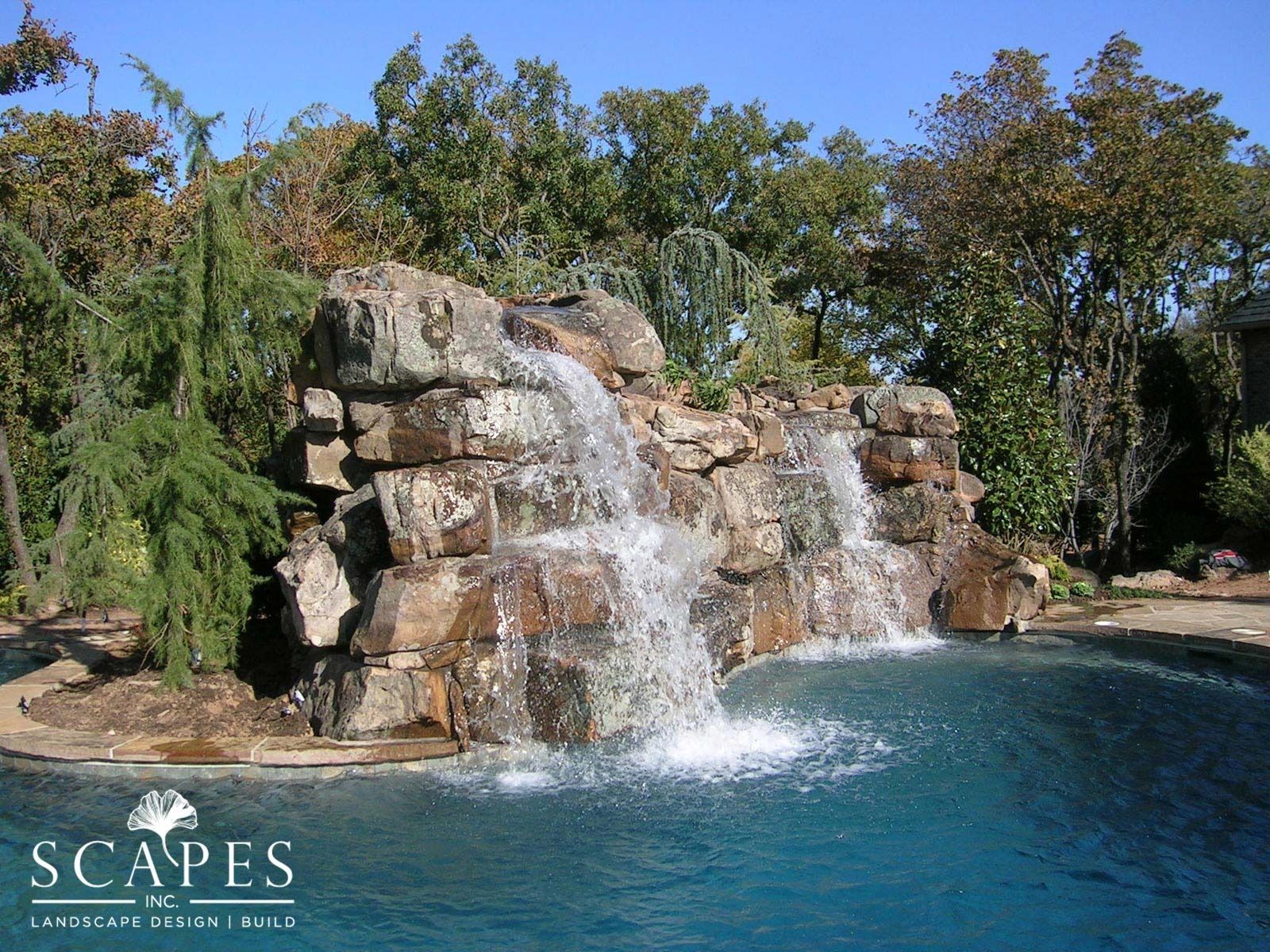 A cascading waterfall flows into a blue swimming pool, surrounded by rock and foliage.