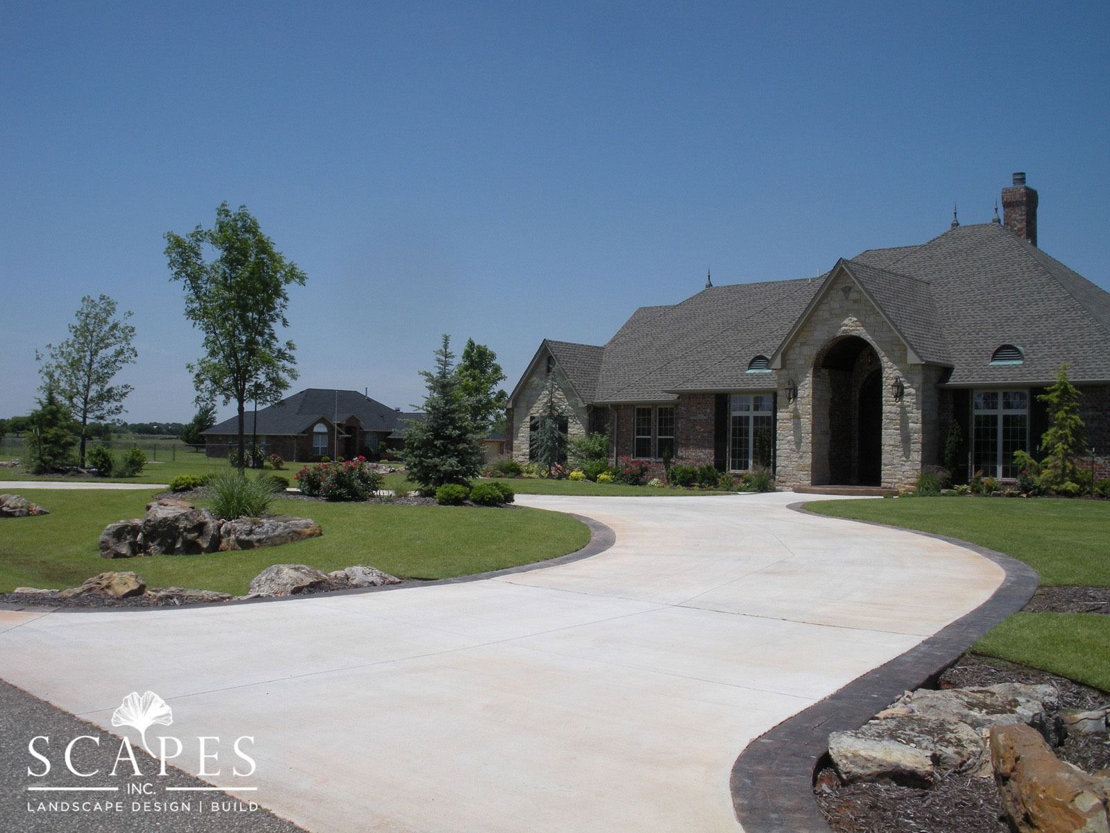 Curving driveway leading to a brick and stone house with landscaped lawn and rocks on a sunny day.
