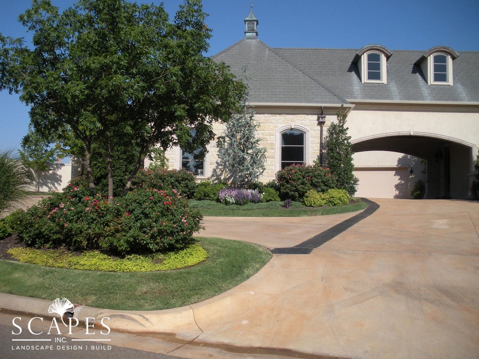Landscaped front yard with a beige house, a circular driveway, and various plants and trees.