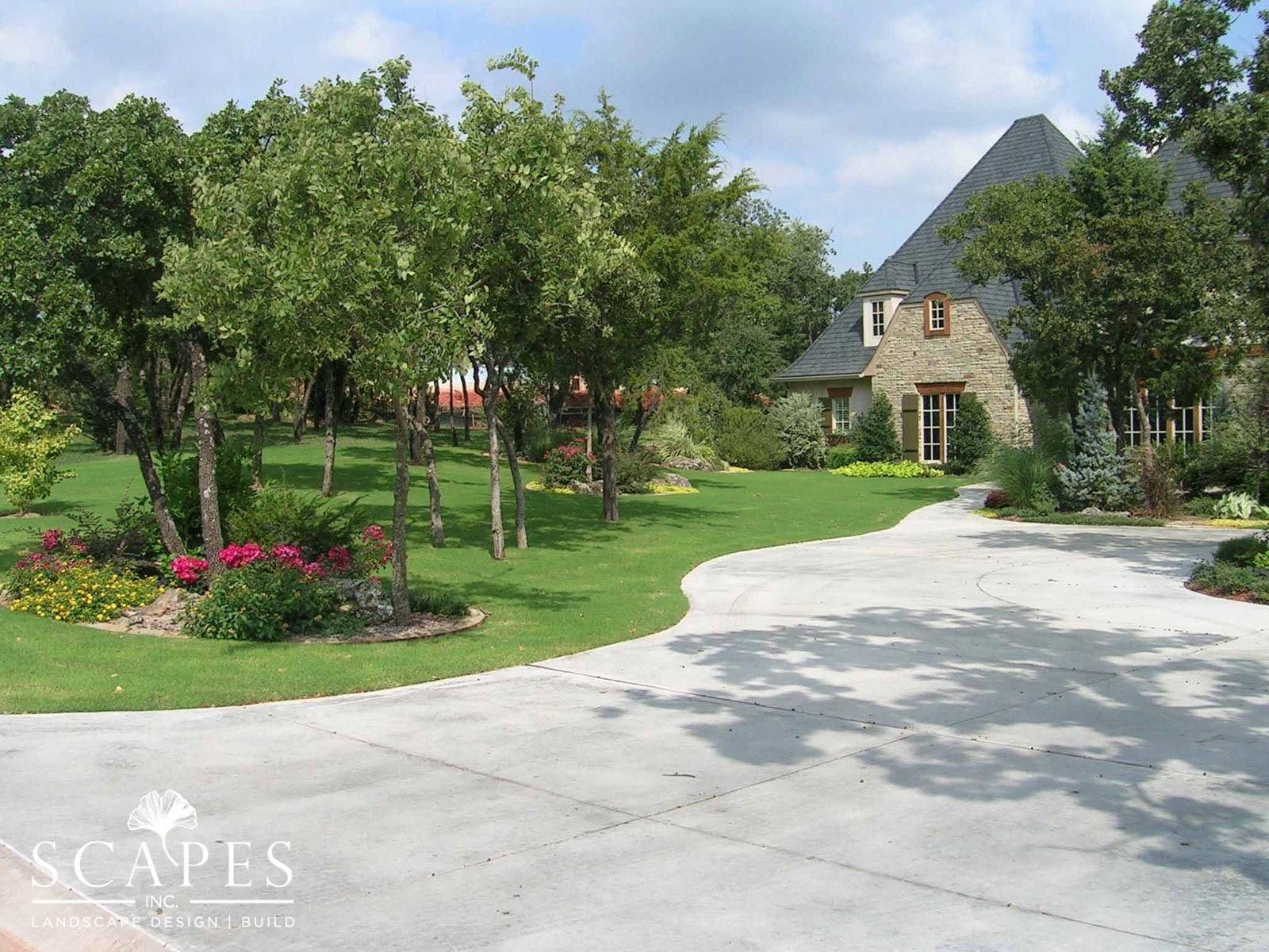 A curved concrete driveway leads to a stone house with a dark roof, surrounded by trees and green grass.
