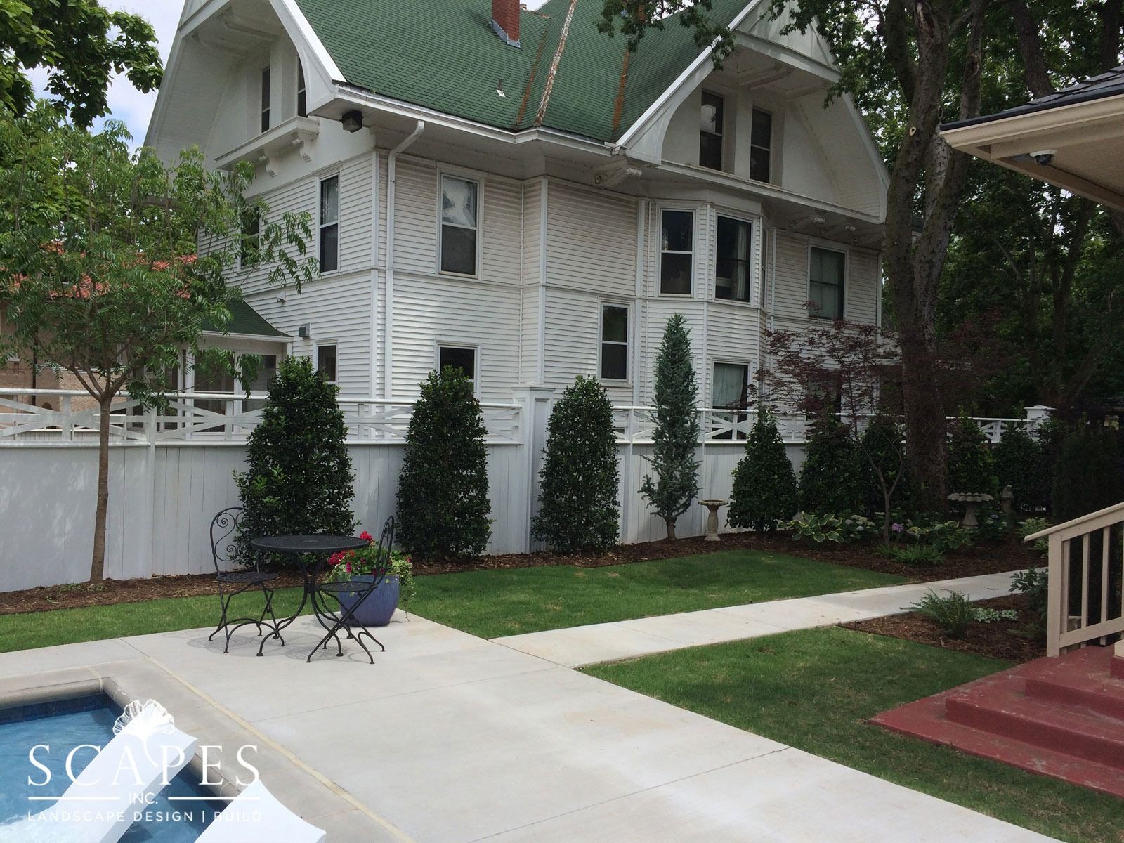 White Victorian house with a green roof, tall evergreen trees, and a patio with a pool.