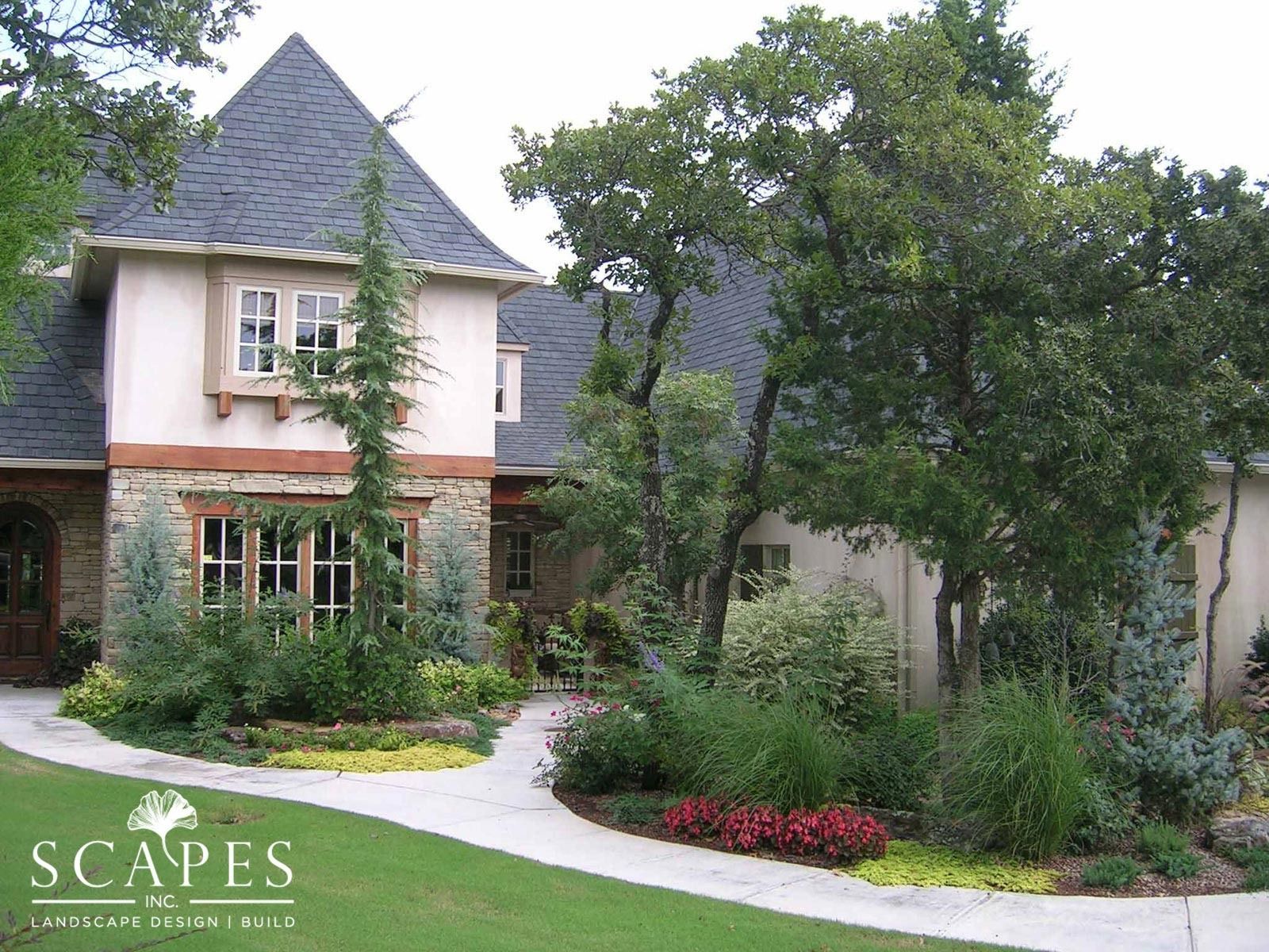 A house with a stone facade and a gray roof, surrounded by landscaping with various green plants and a walkway.